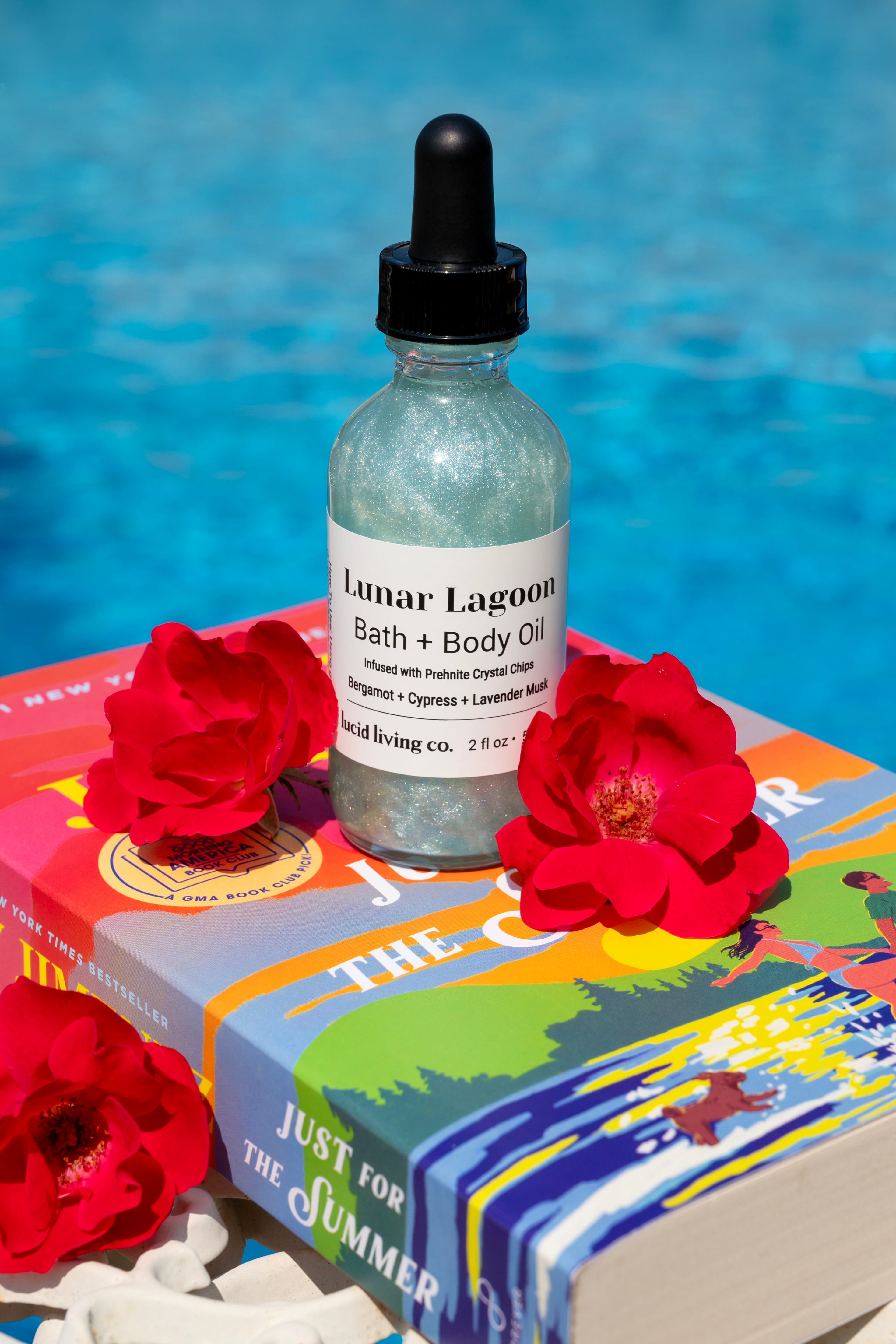 Shimmering bottle of Lunar Lagoon Bath &amp; Body Oil with a black dropper, placed on a colorful summer-themed book by a poolside, surrounded by vibrant red flowers.