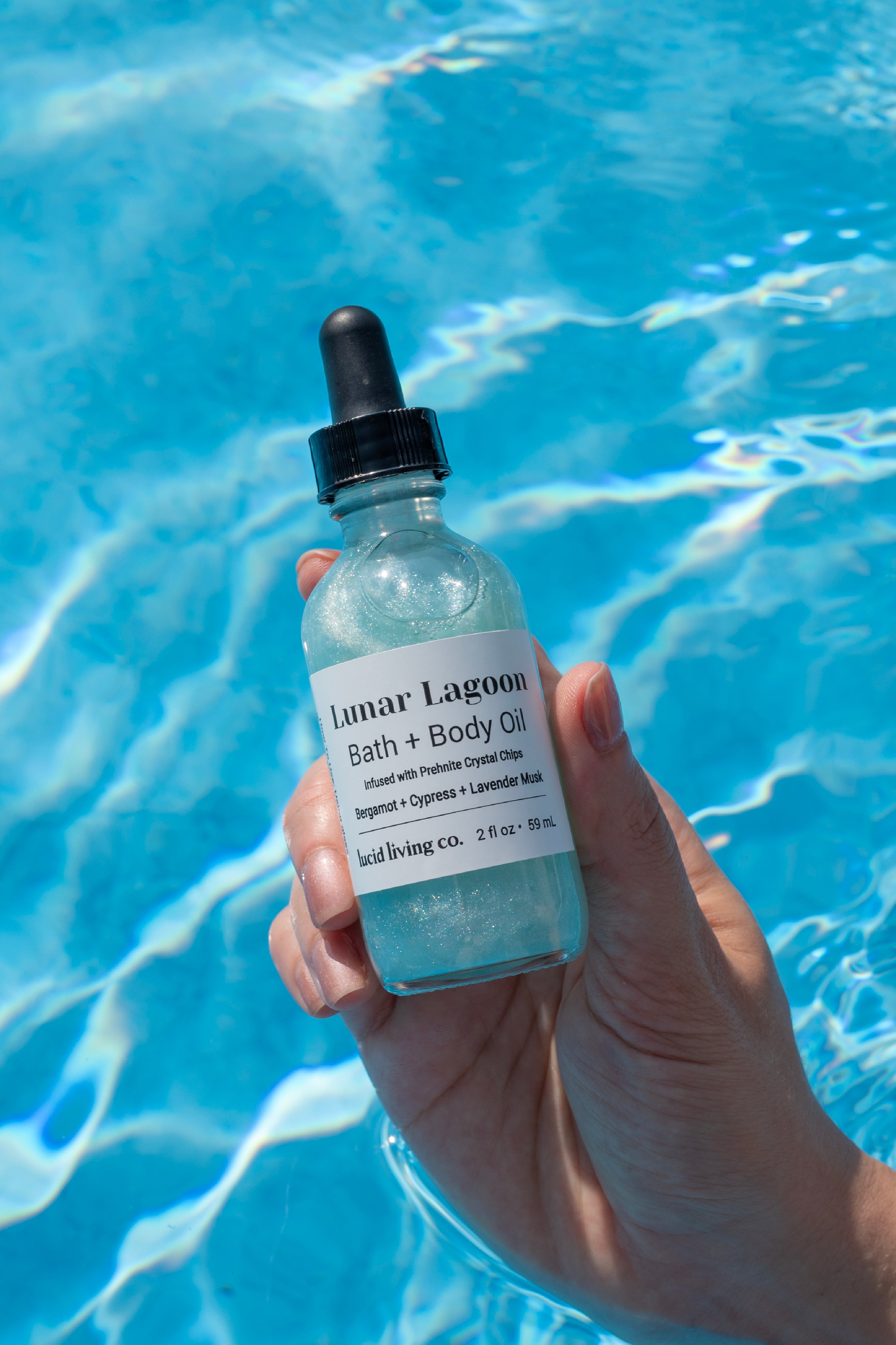 Hand holding a shimmering bottle of Lunar Lagoon Bath &amp; Body Oil with a black dropper, filled with glittering aqua liquid, set against the bright blue rippling water of a pool.