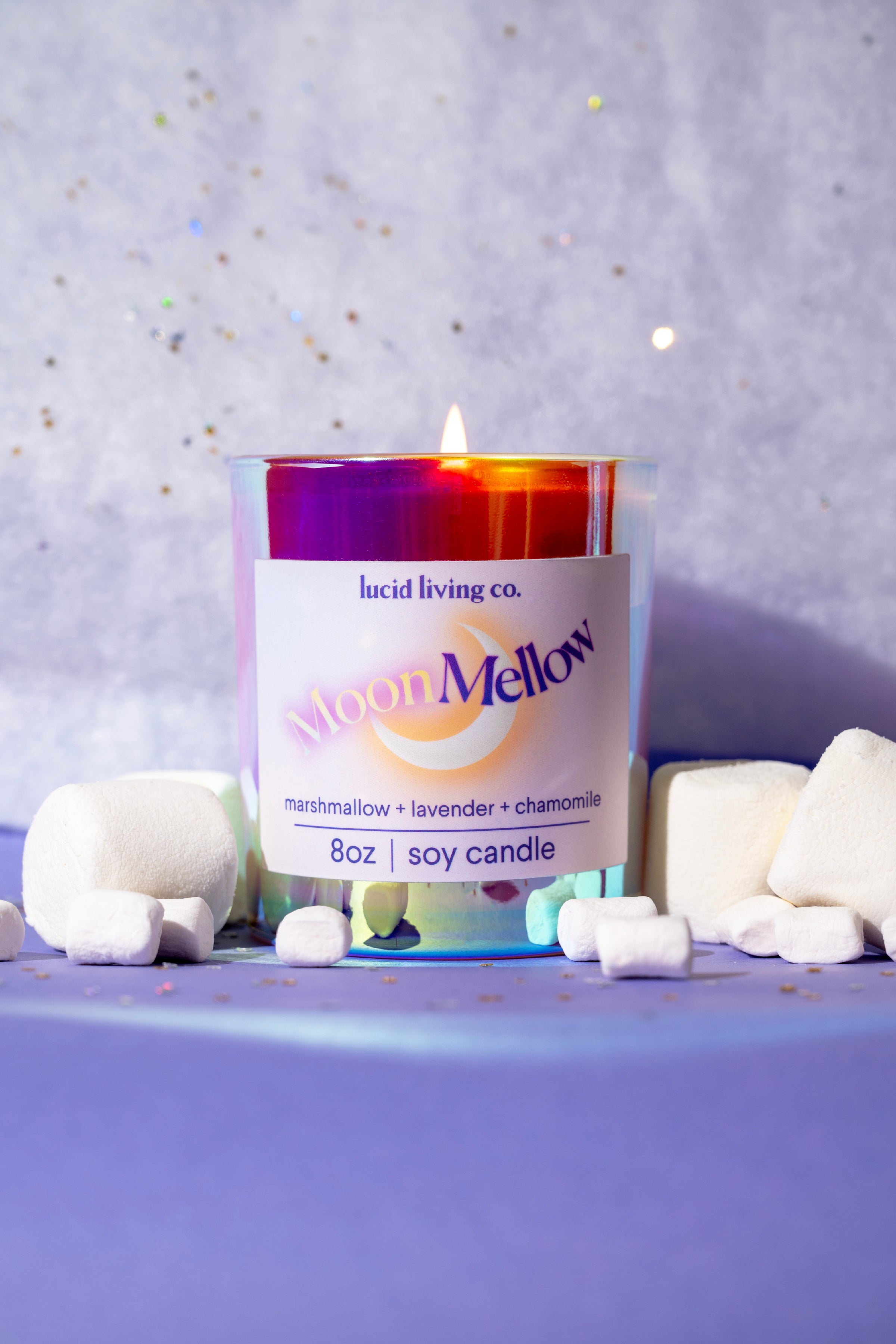 Lit MoonMellow Soy Candle in an iridescent glass jar labeled with marshmallow, lavender, and chamomile scent, surrounded by marshmallows on a lavender background.