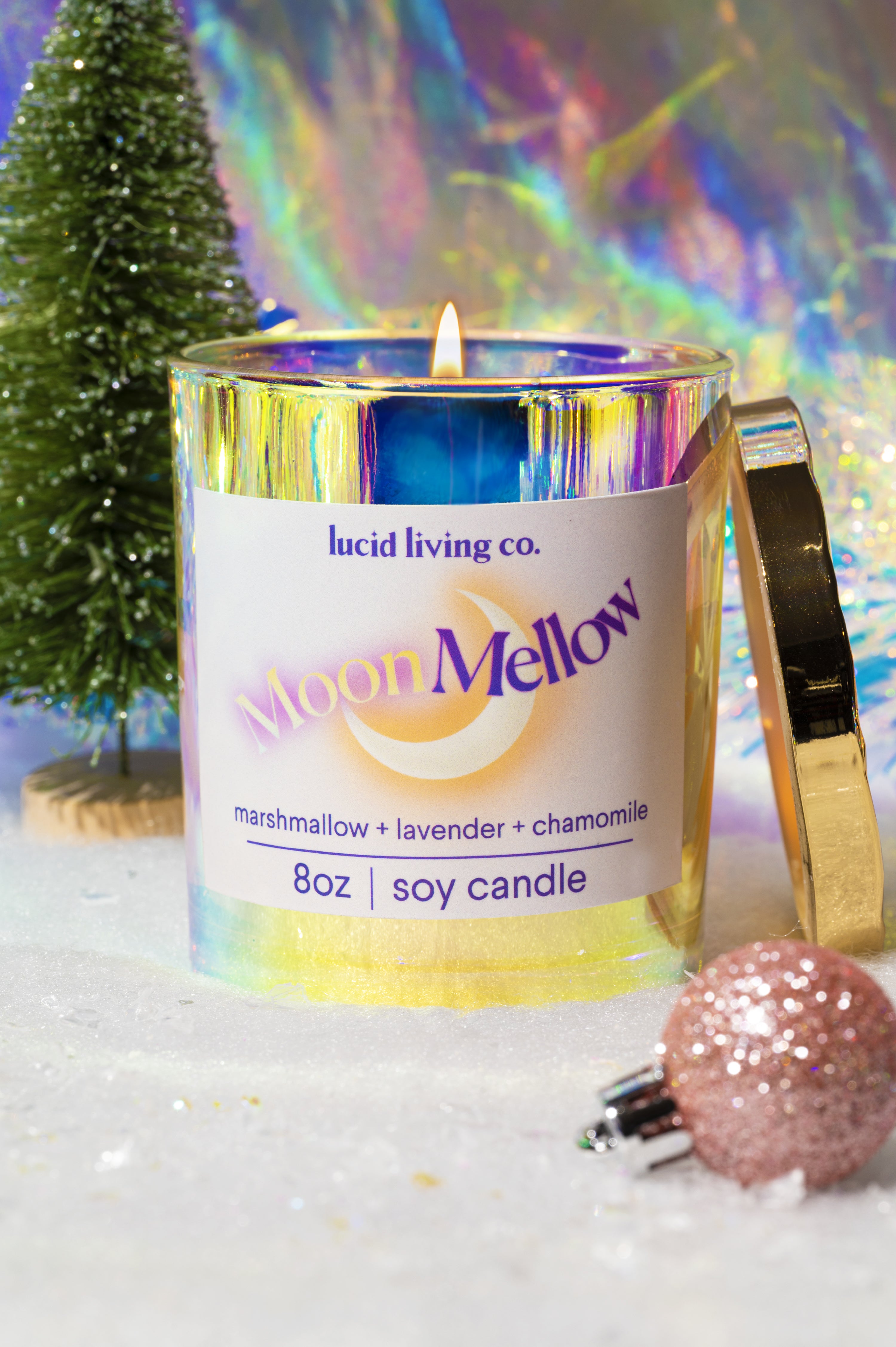 Lit MoonMellow Soy Candle in a reflective glass jar with iridescent colors, placed on a snowy surface decorated with a miniature pine tree and pink glittery ornament.