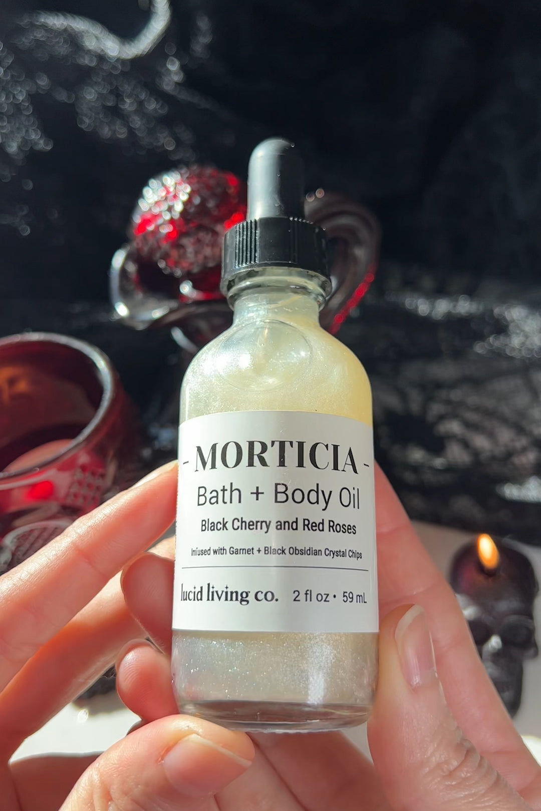 Hand holding a shimmering bottle of MORTICIA Bath &amp; Body Oil with a black dropper cap and white label, glowing softly against a dark, candlelit background.