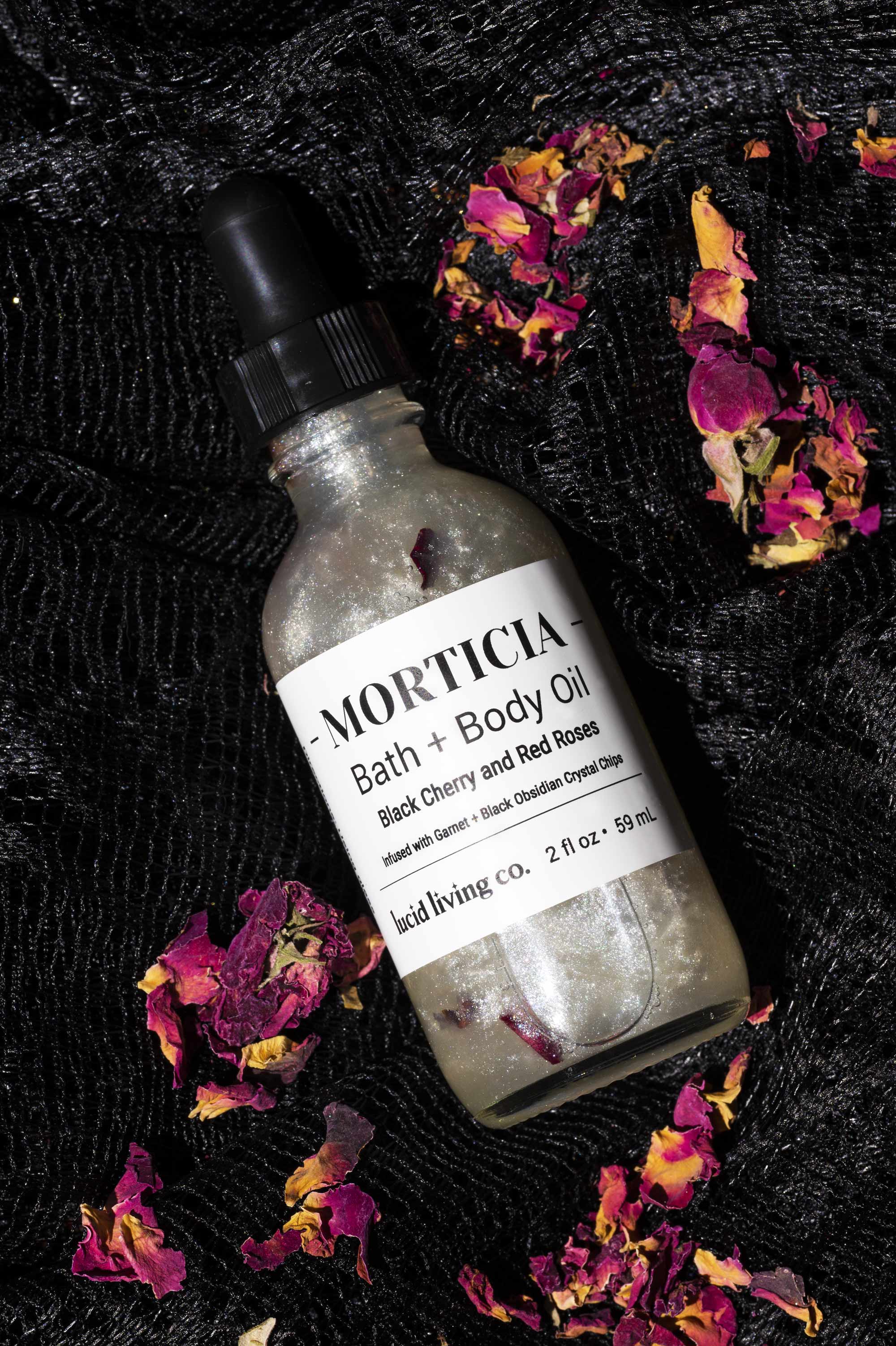 Glass bottle of Morticia Bath + Body Oil with shimmering liquid and rose petals inside, resting on textured black fabric surrounded by dried rose petals, part of the Dark Goddess Bundle.
