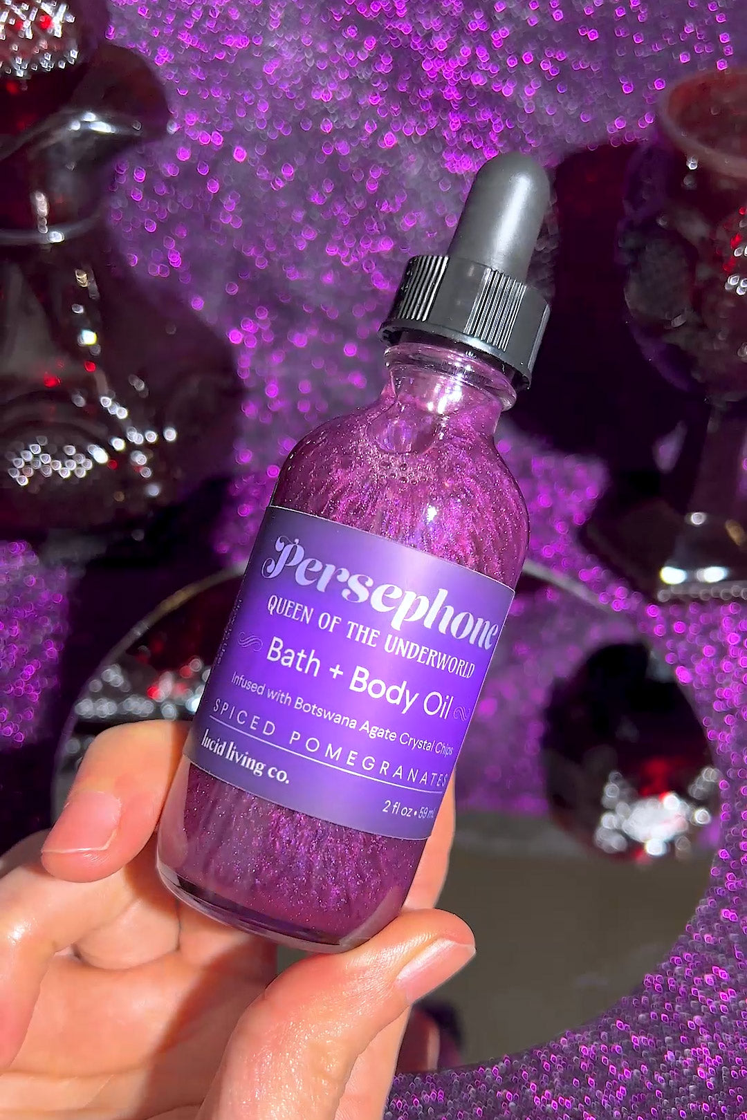 Hand holding a shimmering purple bottle of Persephone Bath + Body Oil with a black dropper, glowing under vibrant purple light and surrounded by reflective, dark decor, part of the Dark Goddess Bundle.