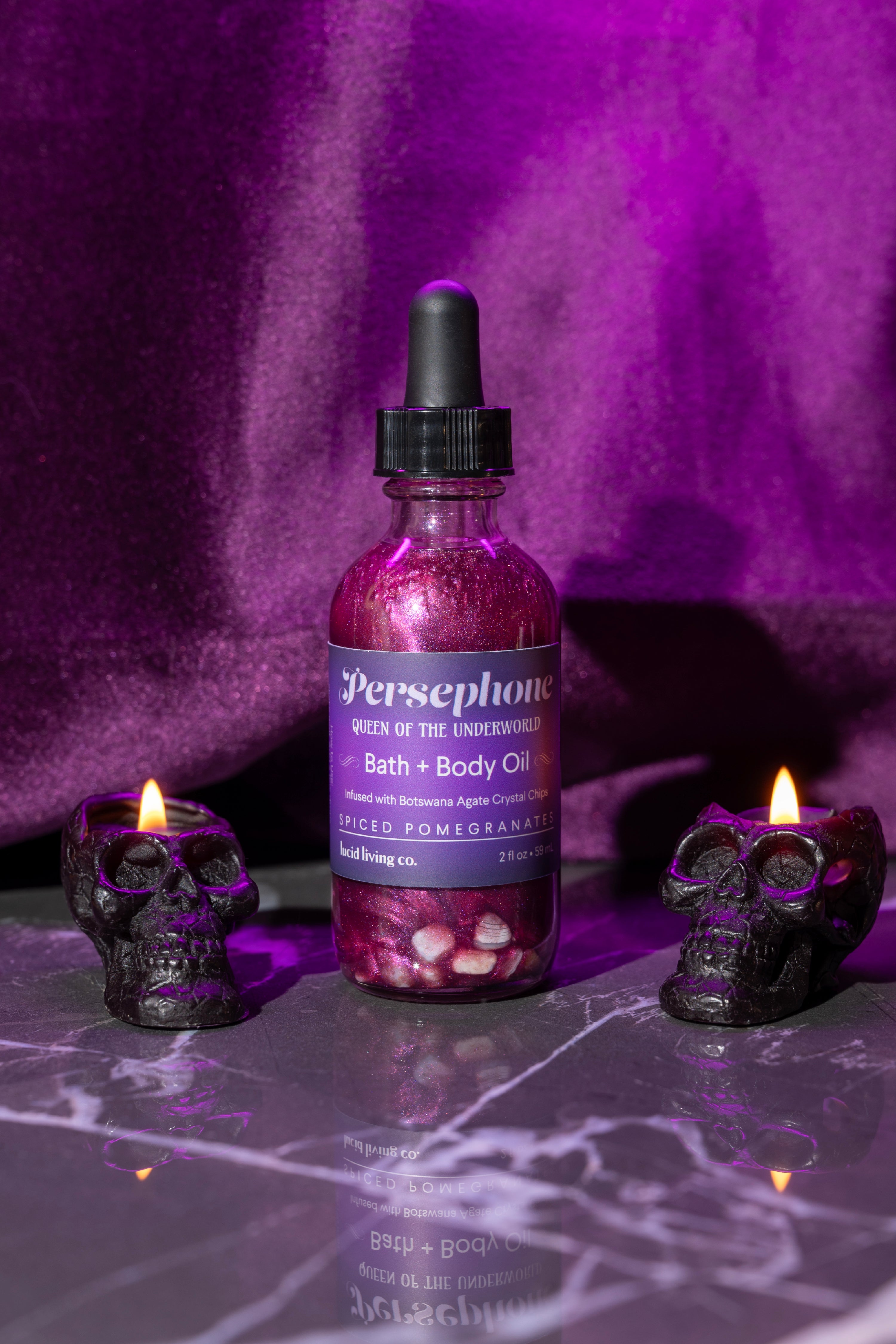 Persephone Bath &amp; Body Oil bottle with shimmering pink liquid and crystal chips, flanked by two black skull-shaped candles with flames on a reflective marble surface.