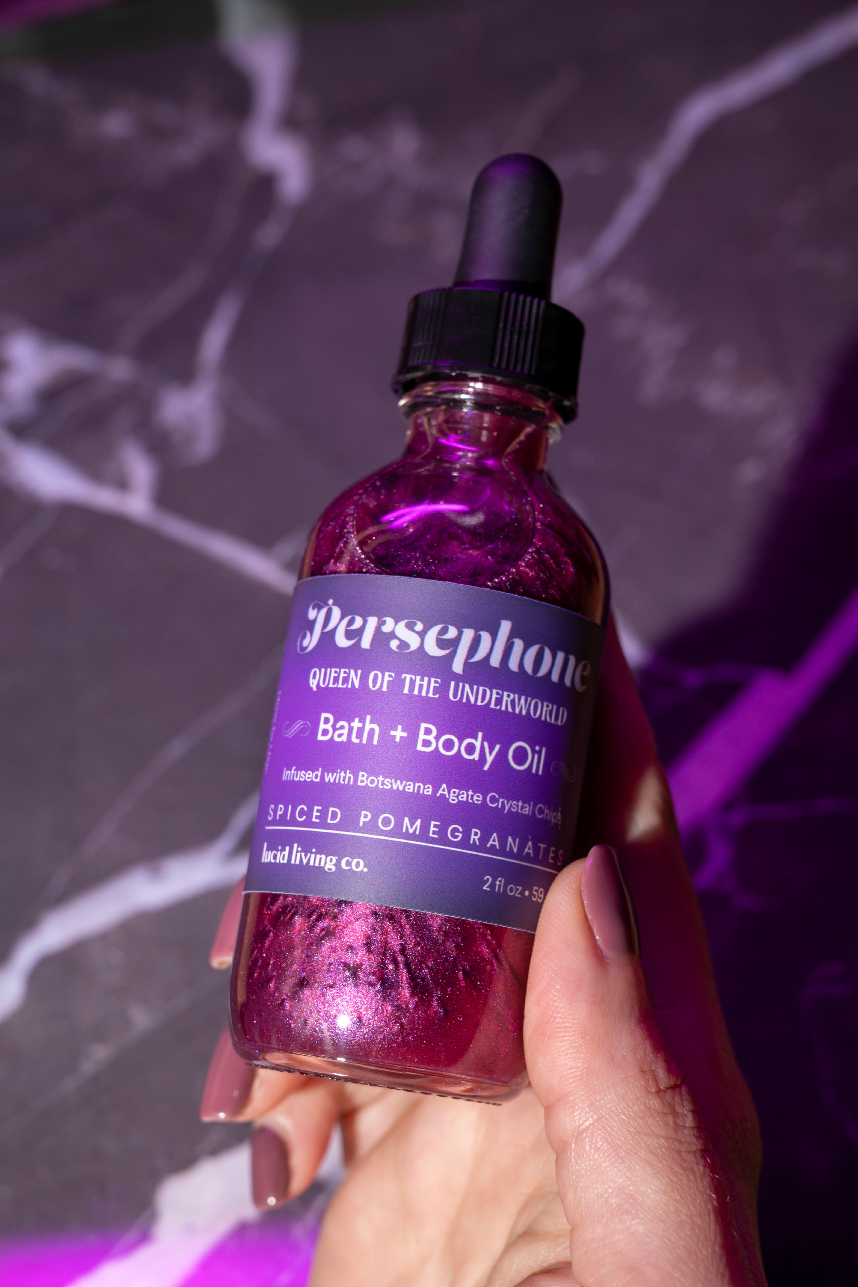 Hand holding a shimmering bottle of Persephone Bath &amp; Body Oil with a deep purple label and black dropper cap, glowing under soft purple lighting on a dark marble surface.