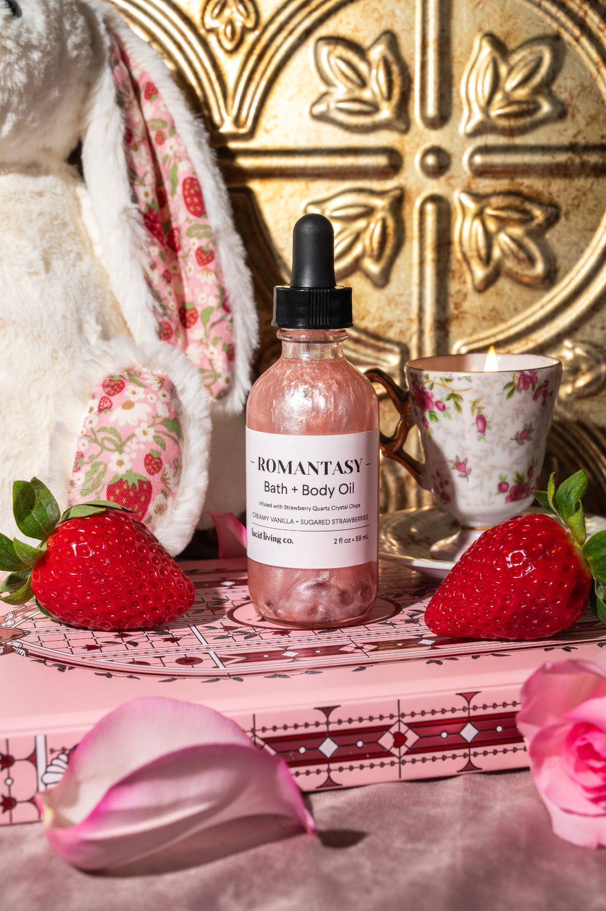 Bottle of Romantasy Bath &amp; Body Oil with shimmering pink liquid and black dropper, surrounded by fresh strawberries, a floral teacup, pink rose petals, and a plush bunny for a cozy, romantic setting.