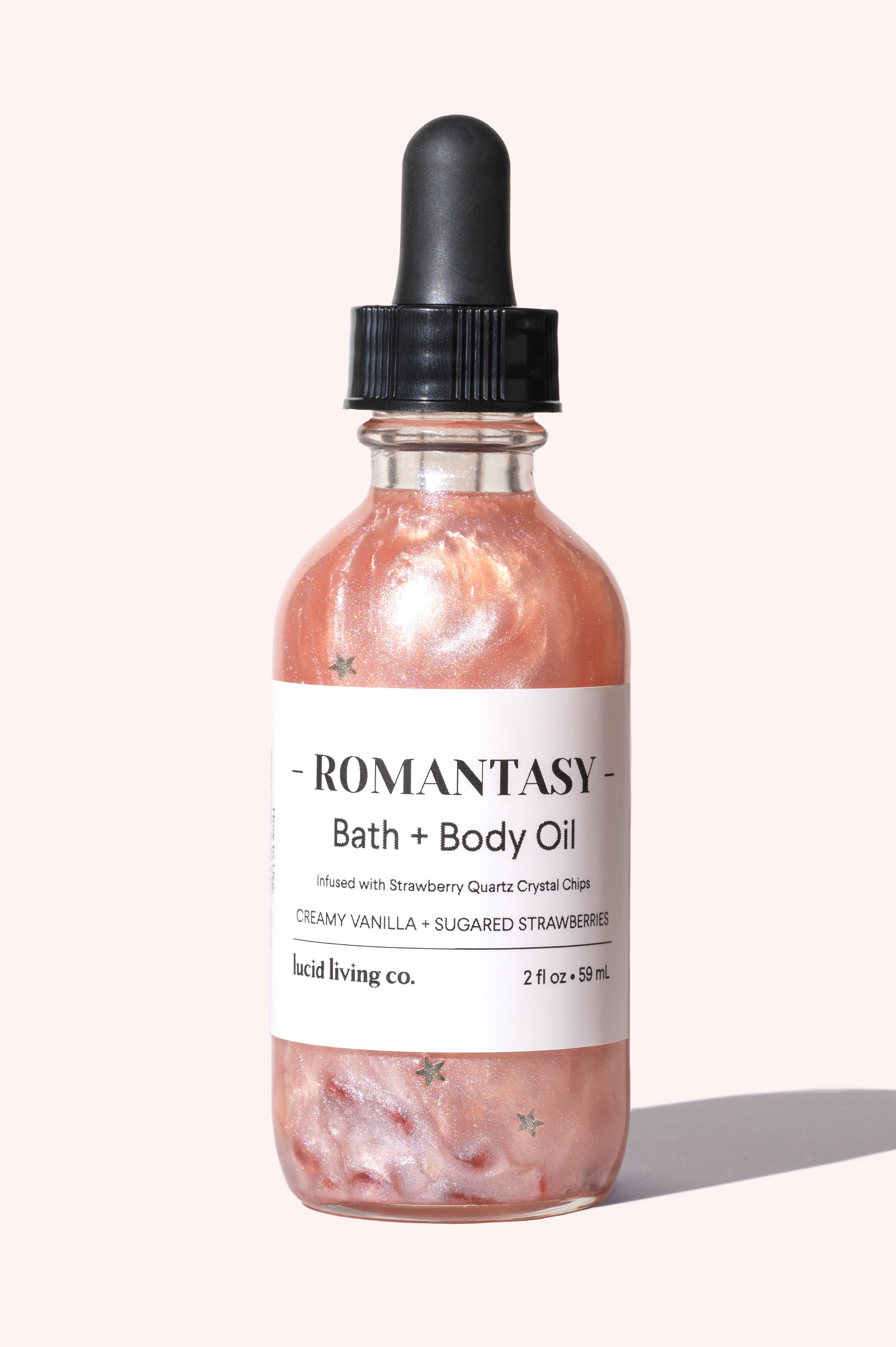 Clear glass bottle of Romantasy Bath &amp; Body Oil with shimmering soft pink liquid and star-shaped flecks, set against a pale pink background highlighting its creamy vanilla and strawberry scent.