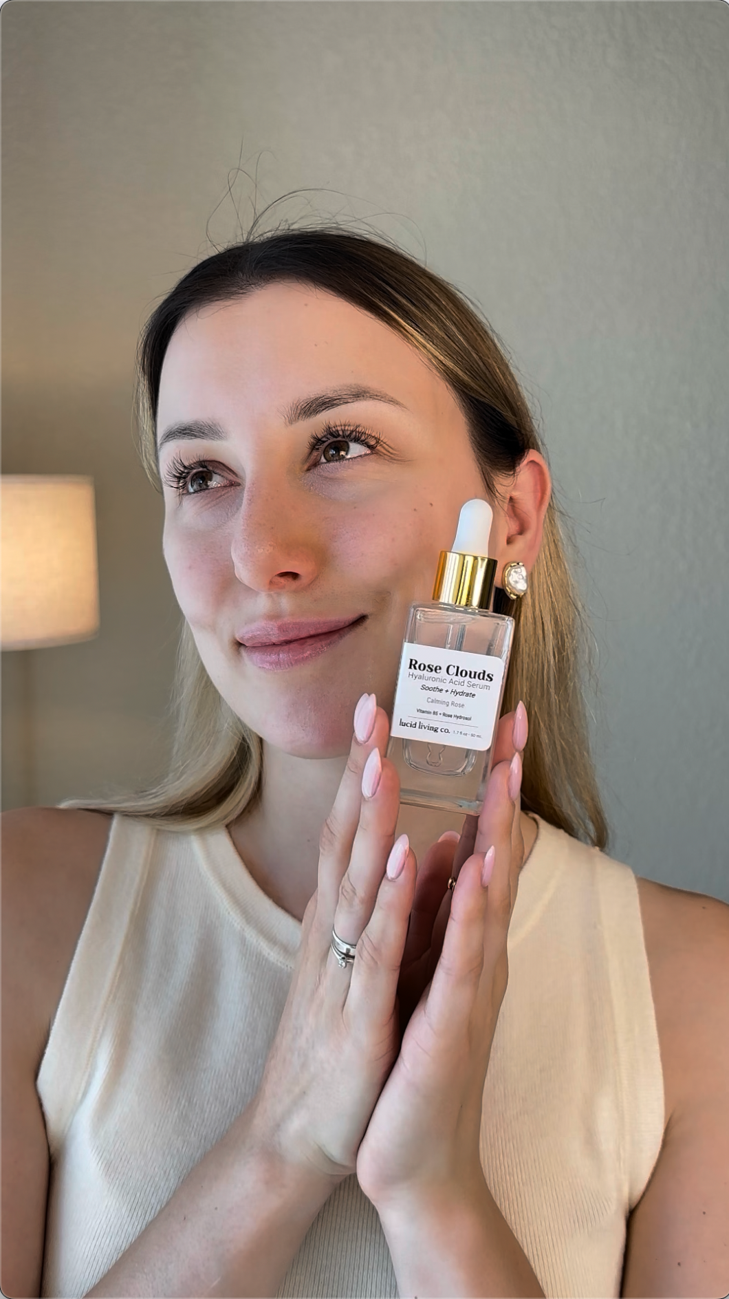 Woman with natural makeup holding a clear glass bottle of Rose Clouds Hyaluronic Acid Serum with a white dropper and gold collar, smiling softly in warm indoor light.