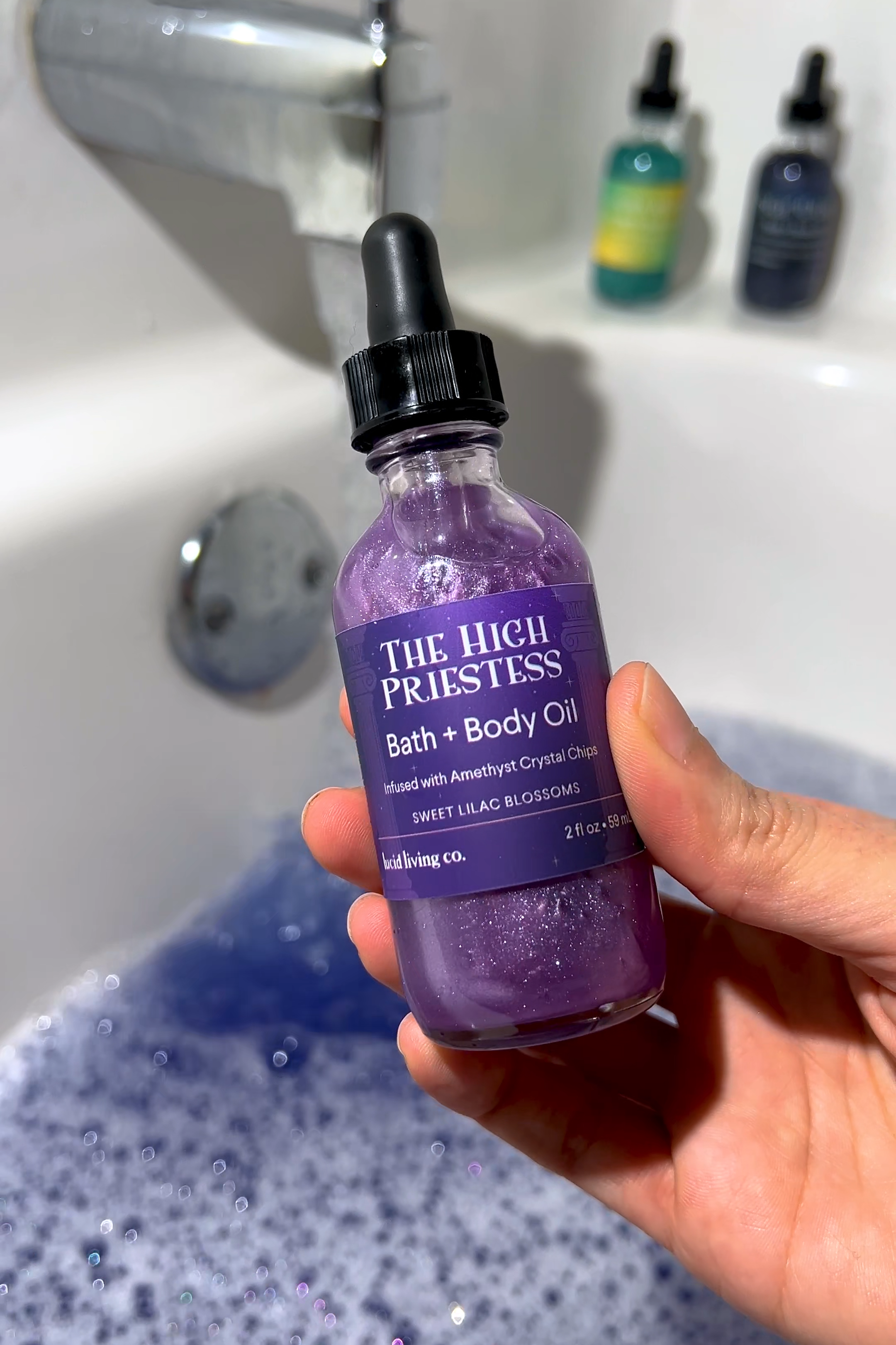 The High Priestess Bath &amp; Body Oil bottle with shimmering purple oil held over a bathtub with bubbles.