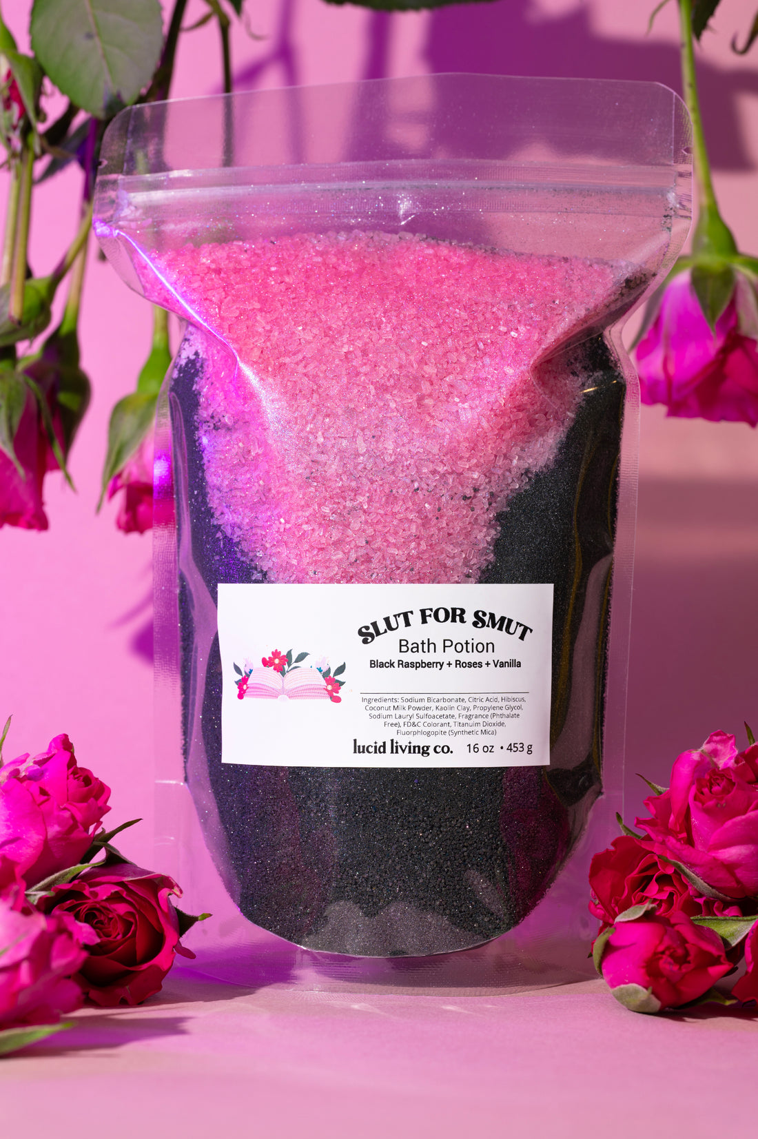 Slut for Smut Bath Potion with black raspberry, roses, and vanilla in a clear bag surrounded by pink roses.