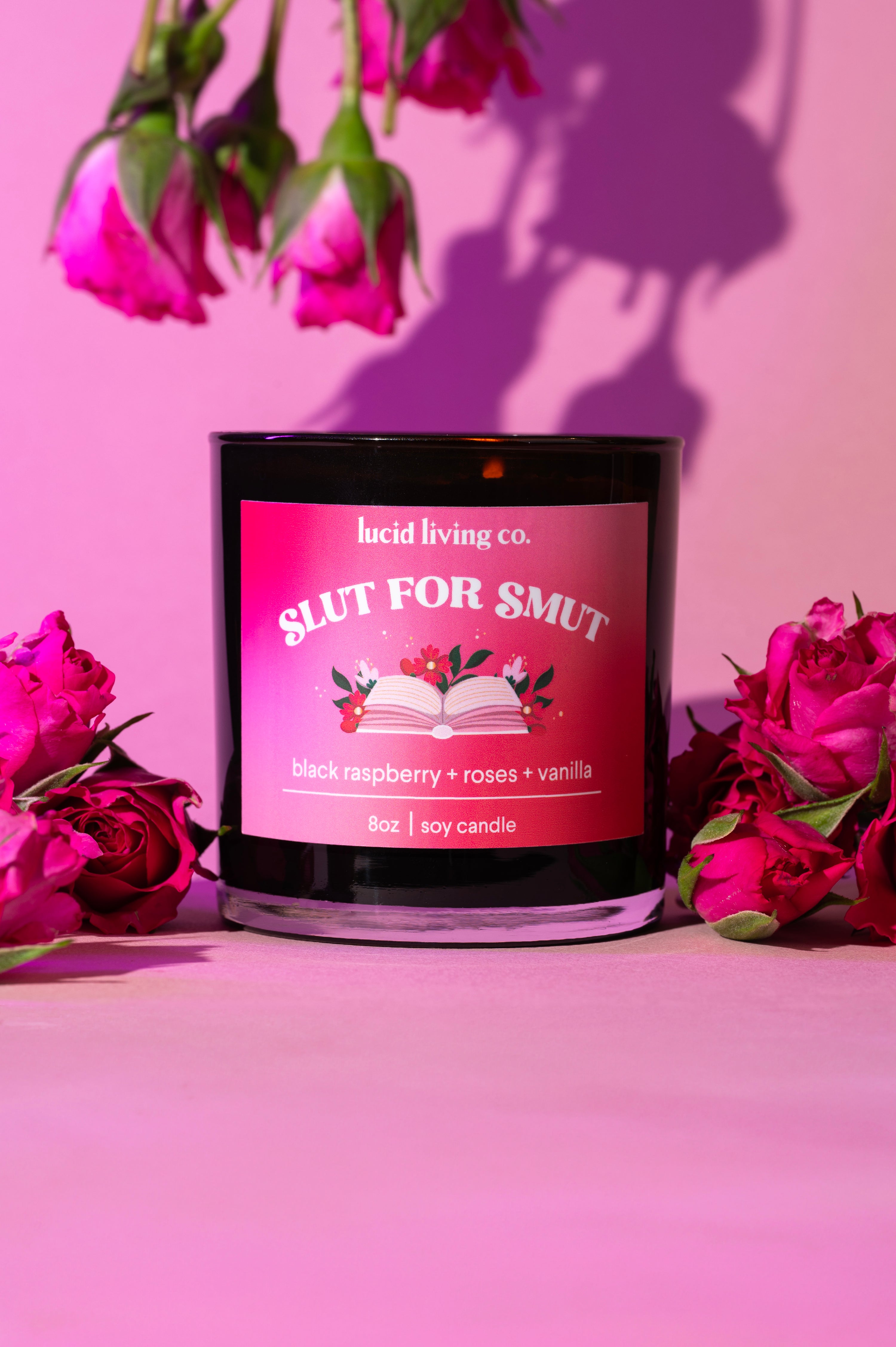 Slut for Smut Soy Candle with black raspberry, roses, and vanilla scent surrounded by pink roses on pink background.