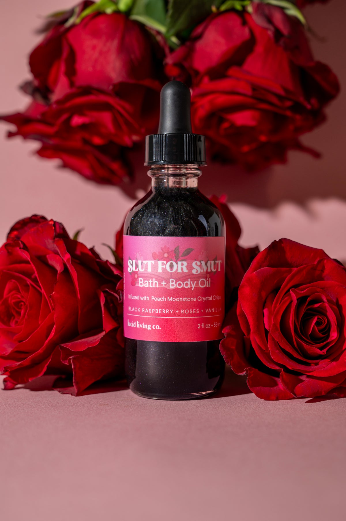 Slut for Smut Bath &amp; Body Oil bottle surrounded by red roses on a pink background.