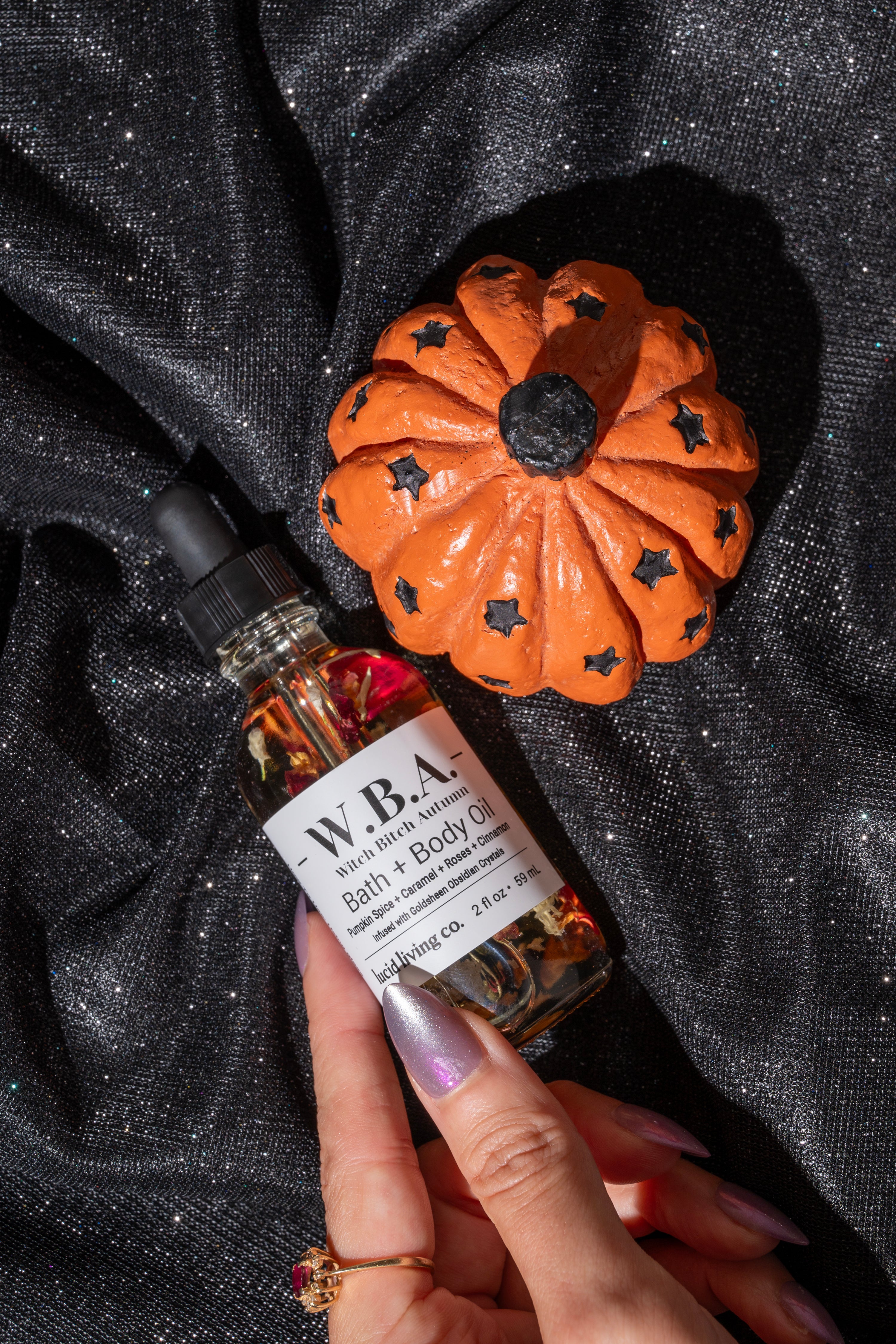 Hand holding a bottle of W.B.A. Bath &amp; Body Oil with a small pumpkin decoration on a black textured background.