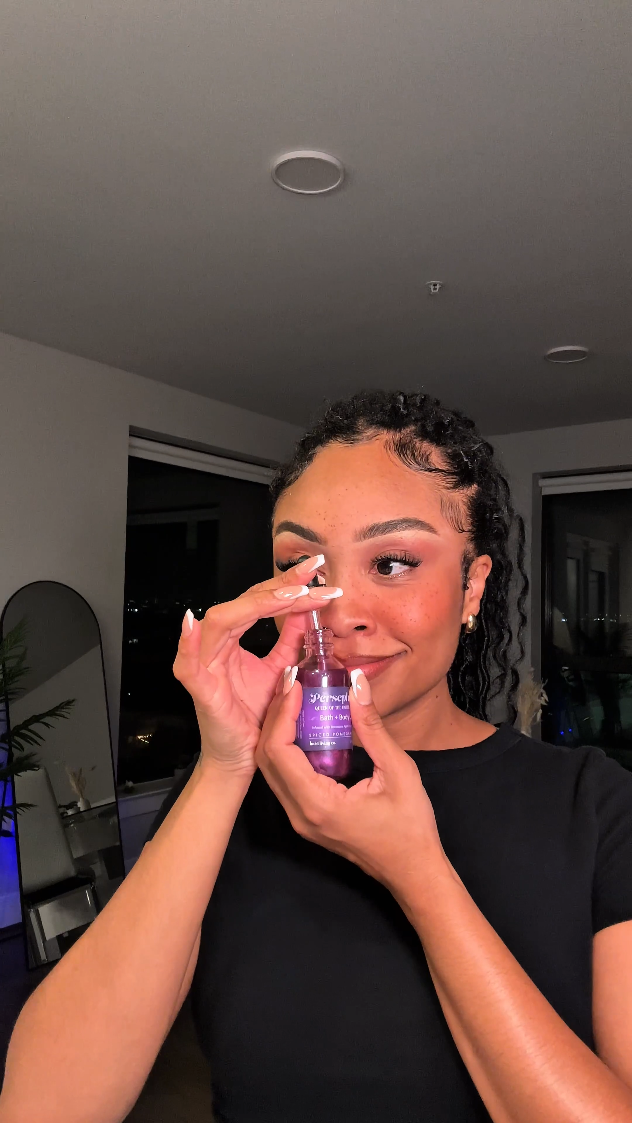 Woman holding and smelling a purple bottle of Persephone Bath & Body Oil indoors, wearing a black top with minimal makeup and a relaxed expression.