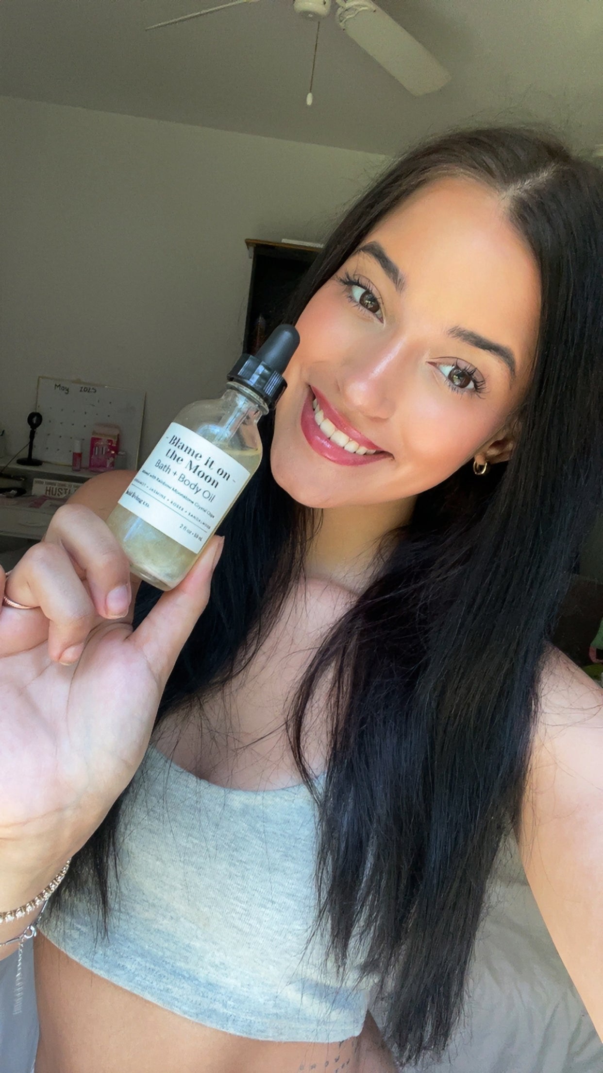 Young woman with long dark hair smiling and holding a clear bottle of Blame it on the Moon Bath & Body Oil with a black dropper in a bright room.