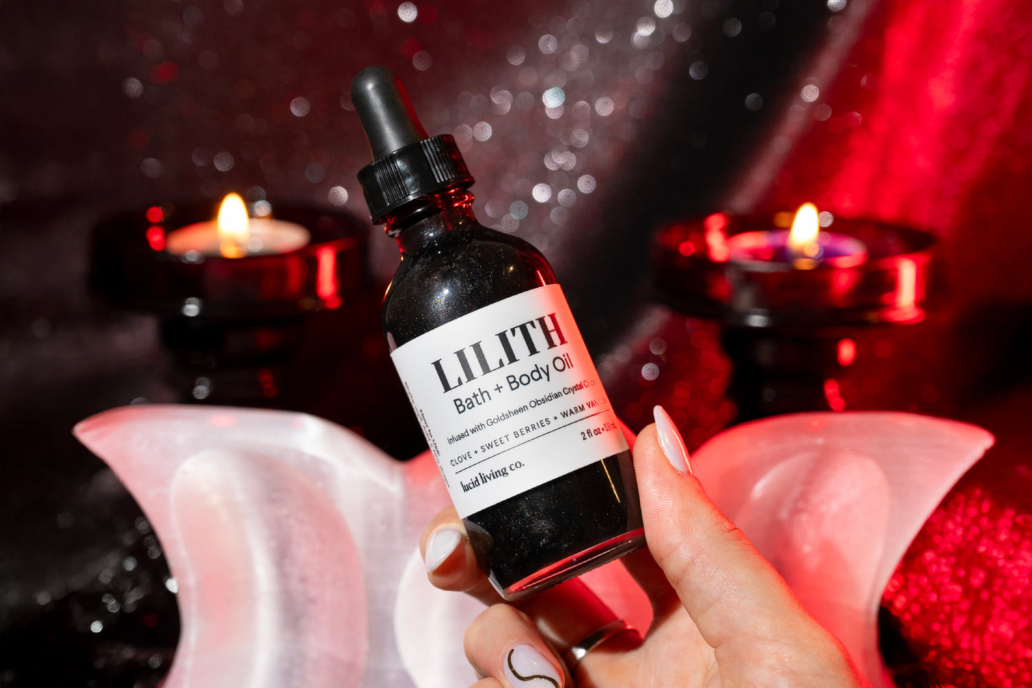 A hand with manicured nails holding a dark glass dropper bottle of bath and body oil labeled “Lilith” against a glittering black and red backdrop with two lit red candles and a pair of crescent-shaped rose quartz crystals.