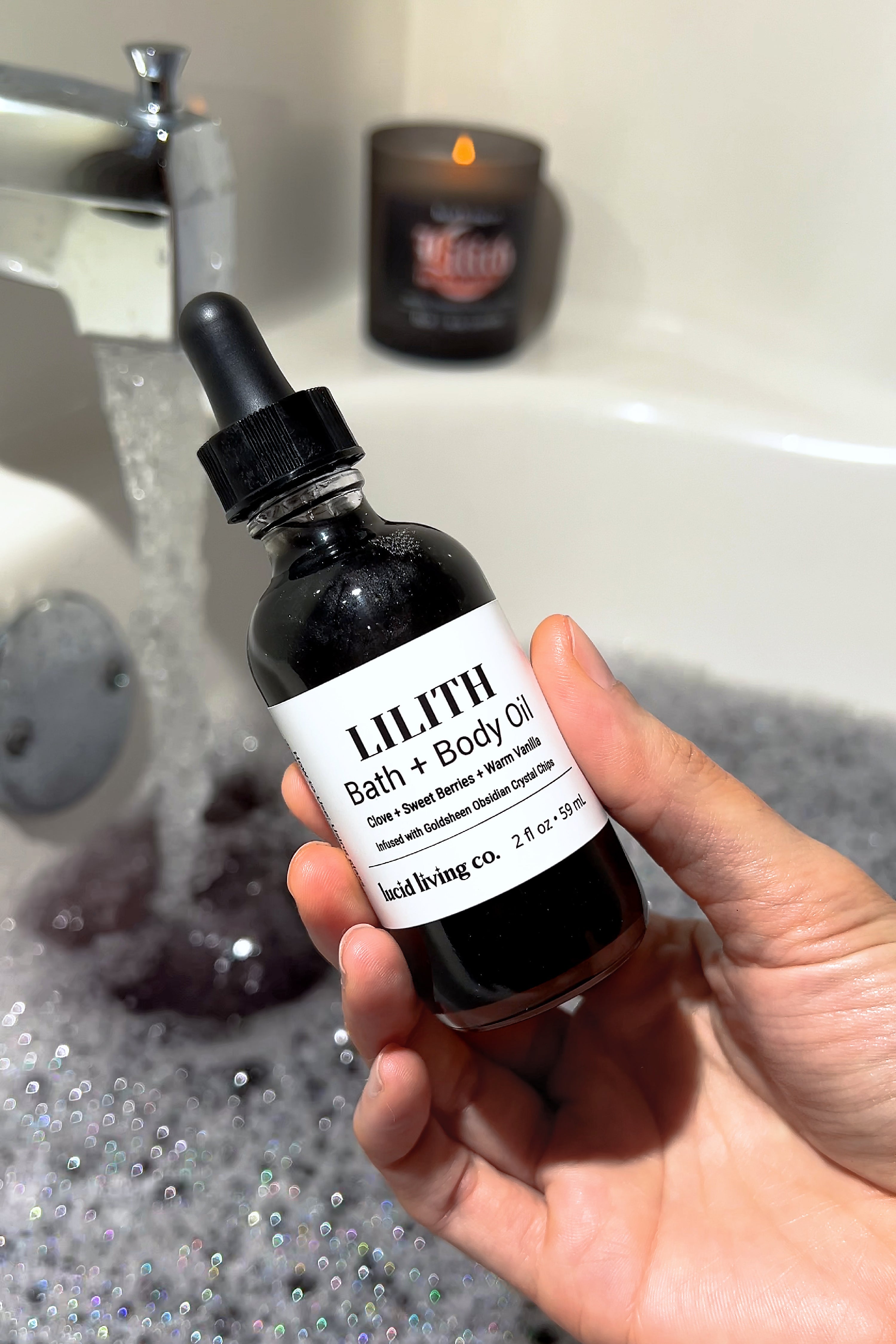LILITH Bath &amp; Body Oil bottle held over a bubbly bathtub with lit candle in the background