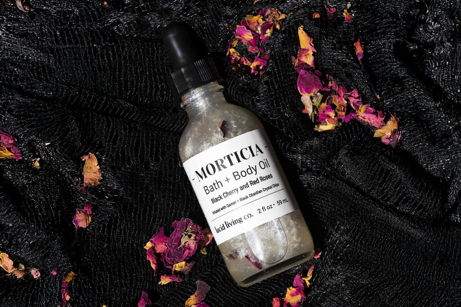 A matte glass bottle of bath and body oil with a black dropper cap labeled with black cherry and red roses scent, resting on black textured fabric with scattered dried rose petals.