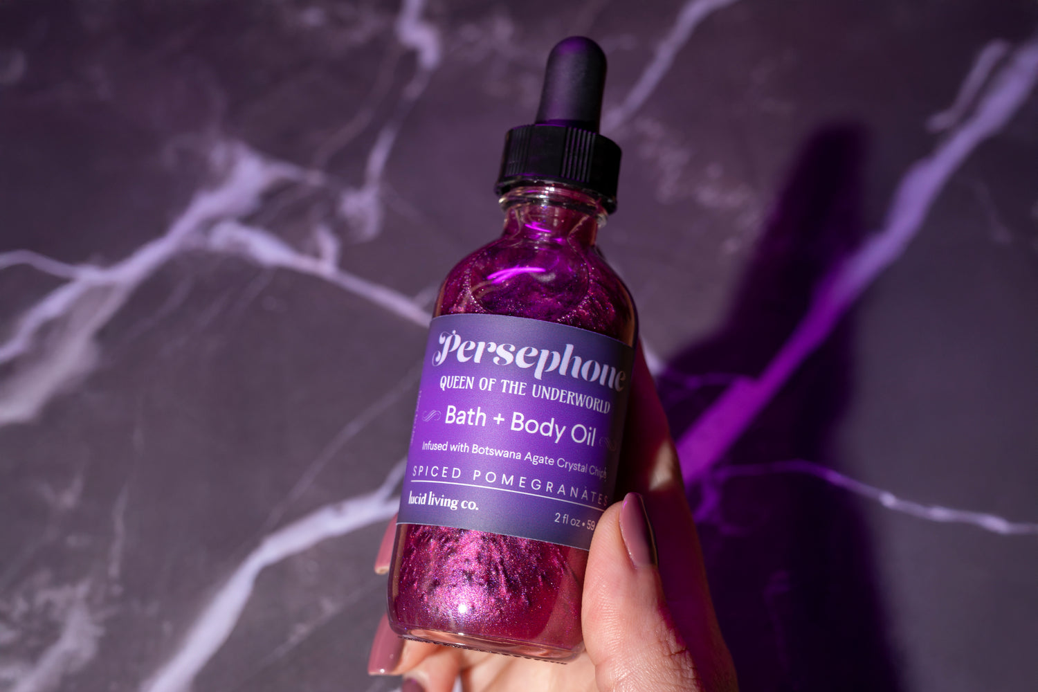 A hand with natural nails holds a purple glass bottle of bath and body oil labeled with a spiced pomegranate scent, set against a dark marbled surface with purple lighting.