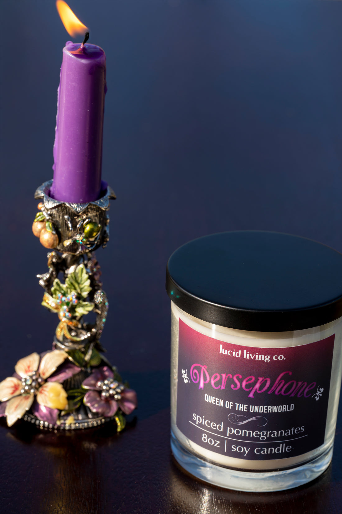 Persephone Soy Candle in a black jar beside a lit purple candle in an ornate floral holder.