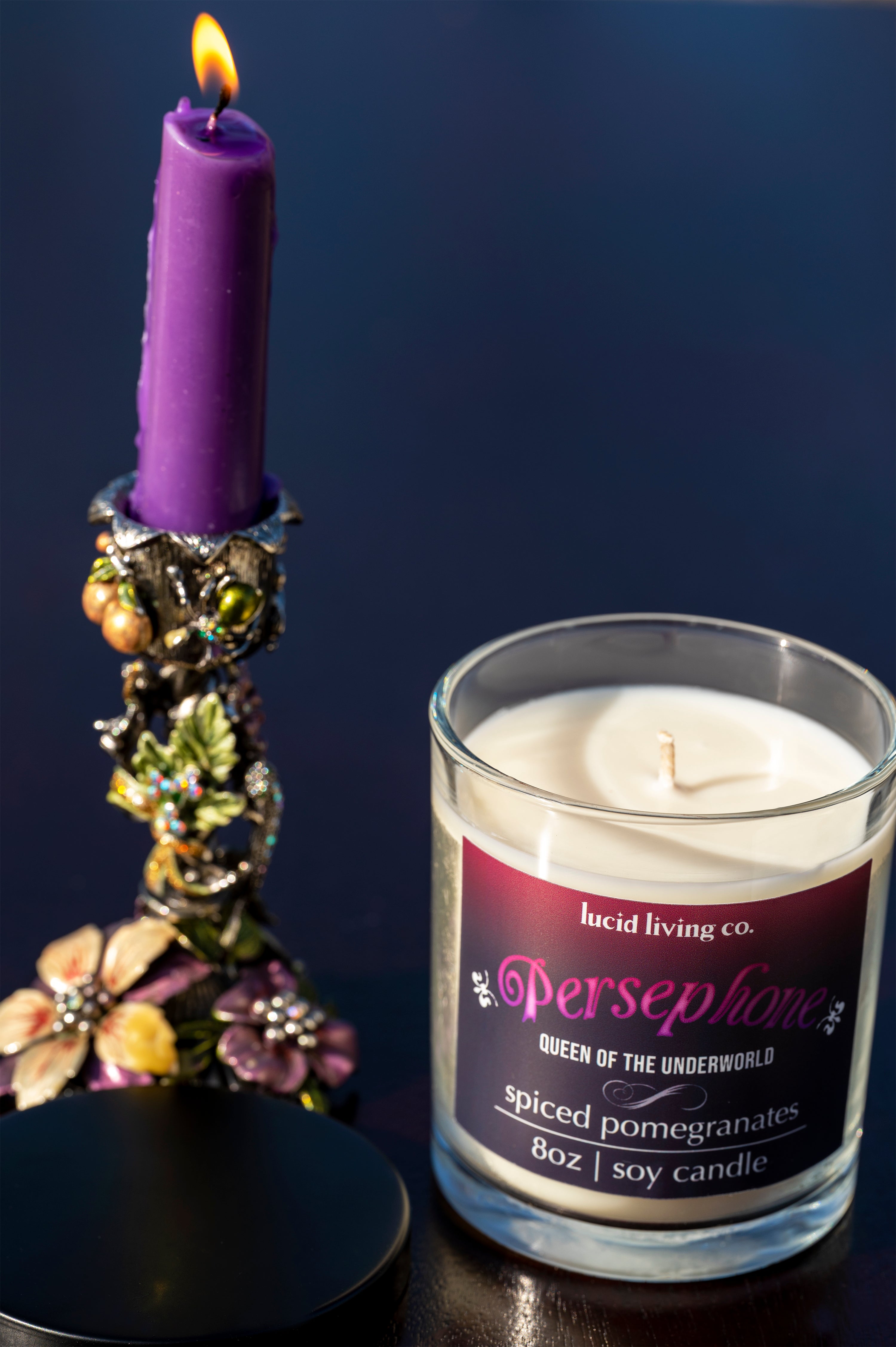 Persephone Soy Candle in clear glass jar with spiced pomegranate scent next to a lit purple candle in floral holder